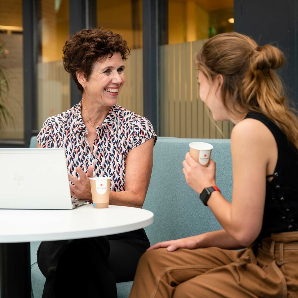 Twee zittende vrouwen lachend in gesprek onder het genot van een kopje koffie.